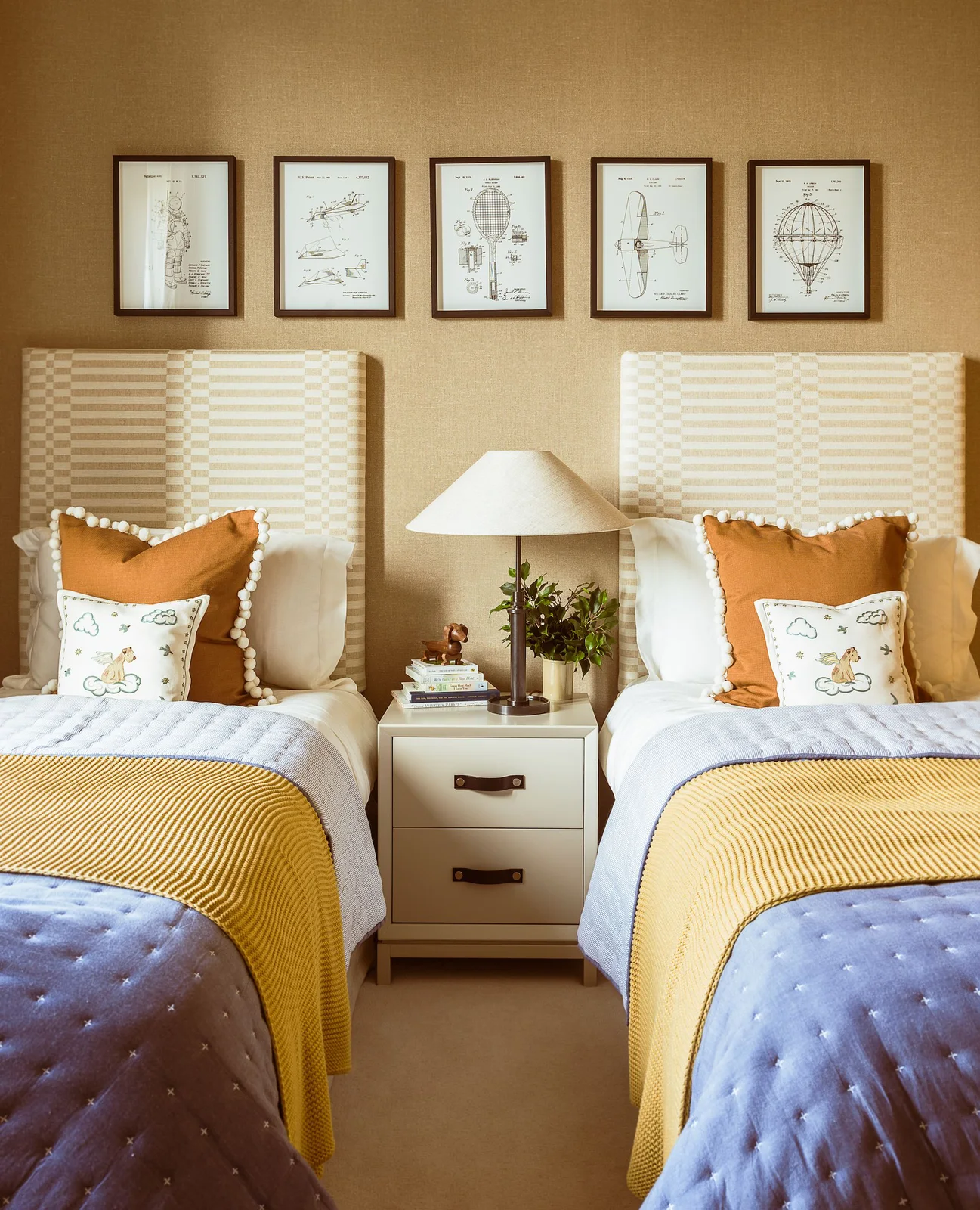 Two pale upholstered beds have blue quilts and yellow knitted throws against a muted golden wall with a central white chest and lamp.