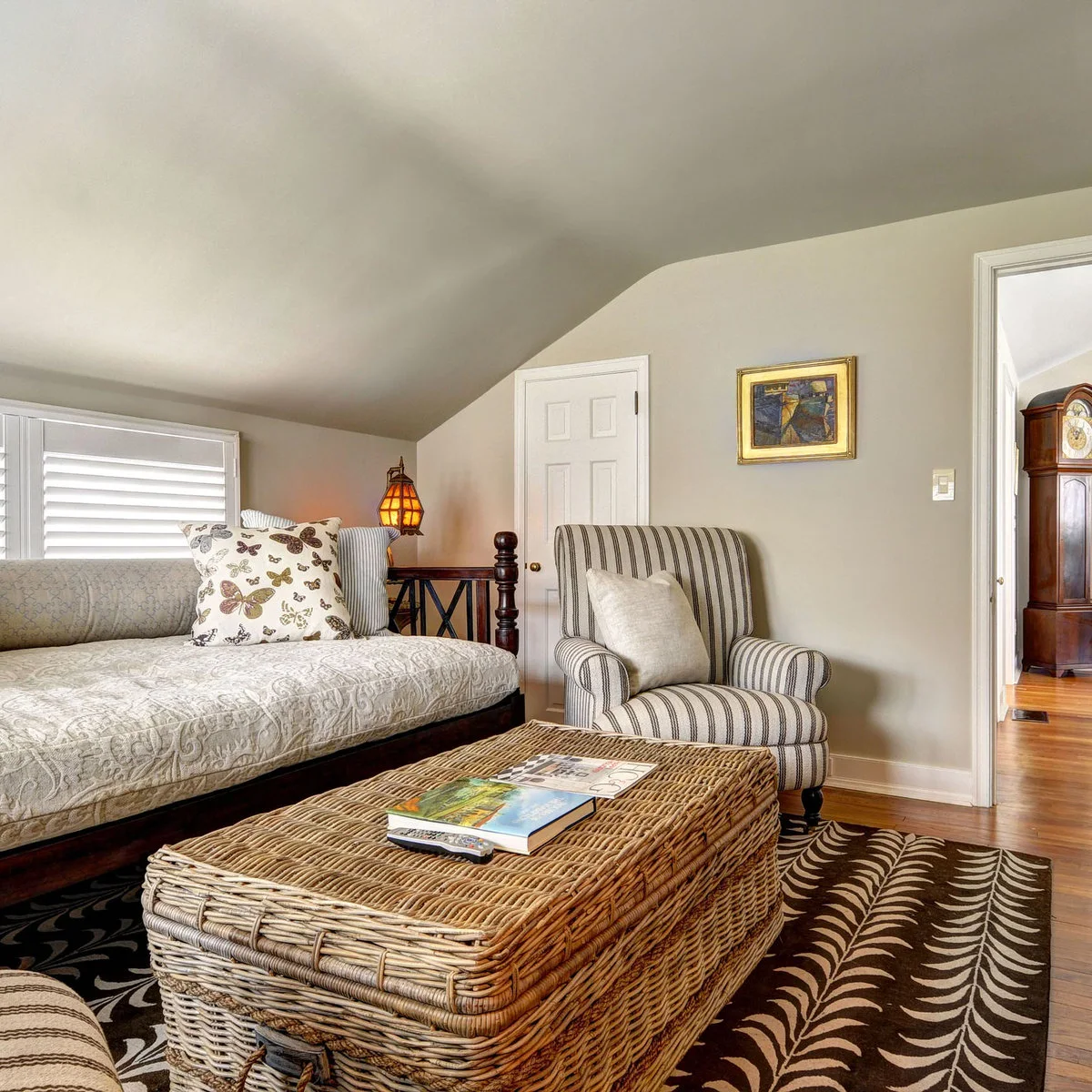 A dark-framed daybed and striped armchair sit under a sloping ceiling around a broad woven-looking ottoman.