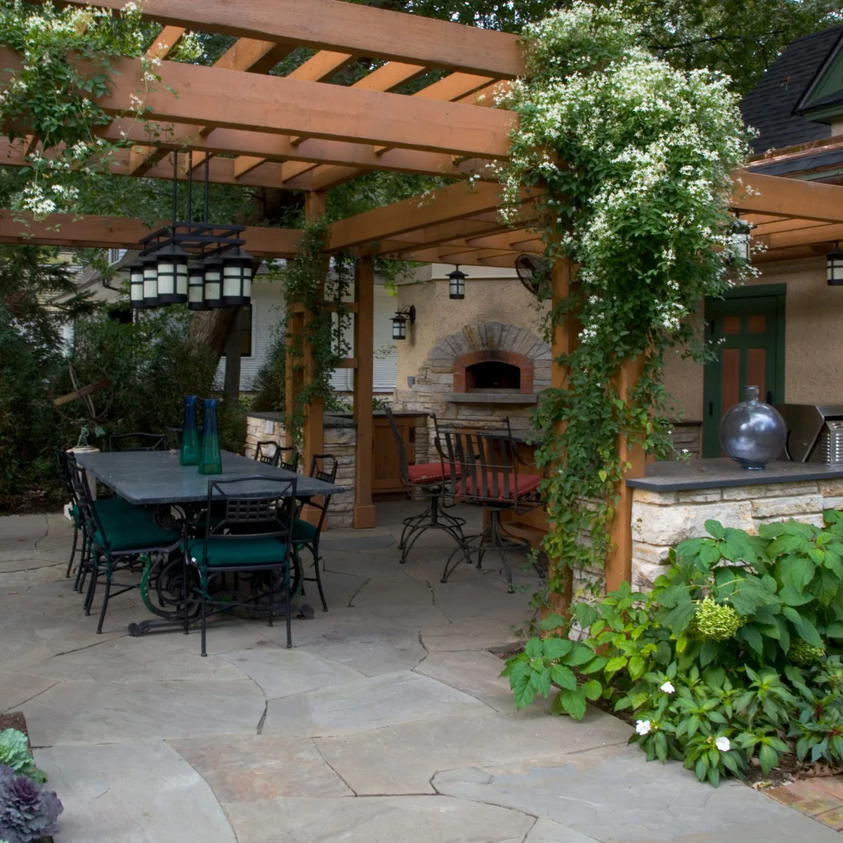 Stone-faced garden bar with dark metal stools beneath a timber pergola covered in climbing greenery and pale flowers