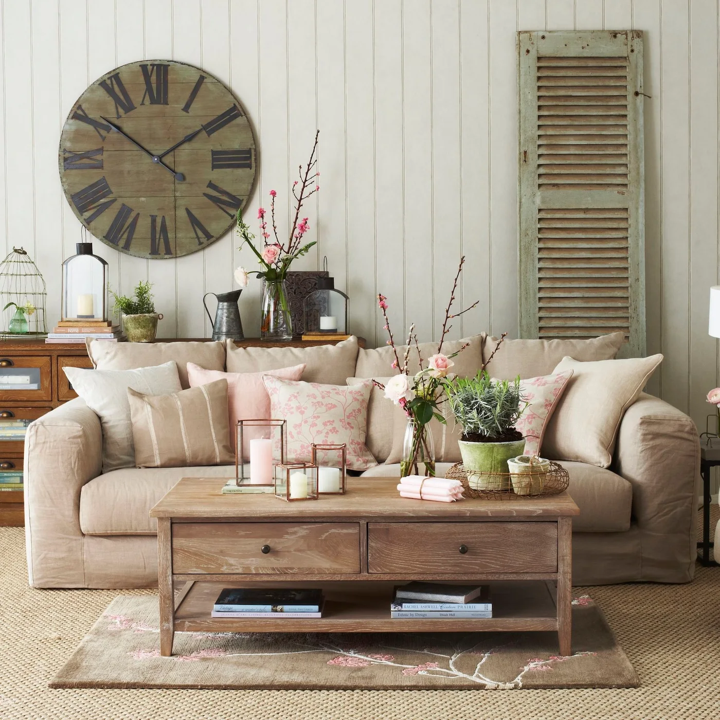A pale sofa sits behind a wooden drawer coffee table below a large clock and beside a weathered shutter-style panel