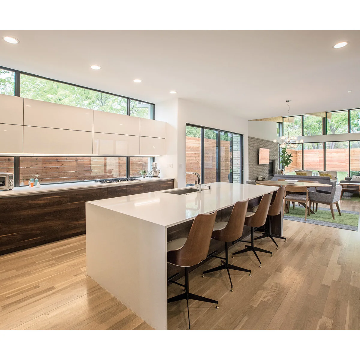 A pale island with brown stools connects a wood-and-white kitchen to a dining table and glazed living area.