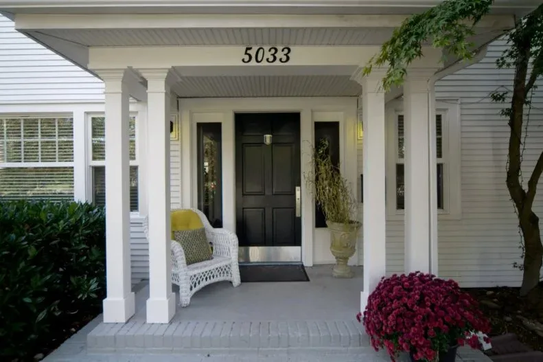 Compact white porch with black door, white woven chair, tall pale planter and visible house numbers
