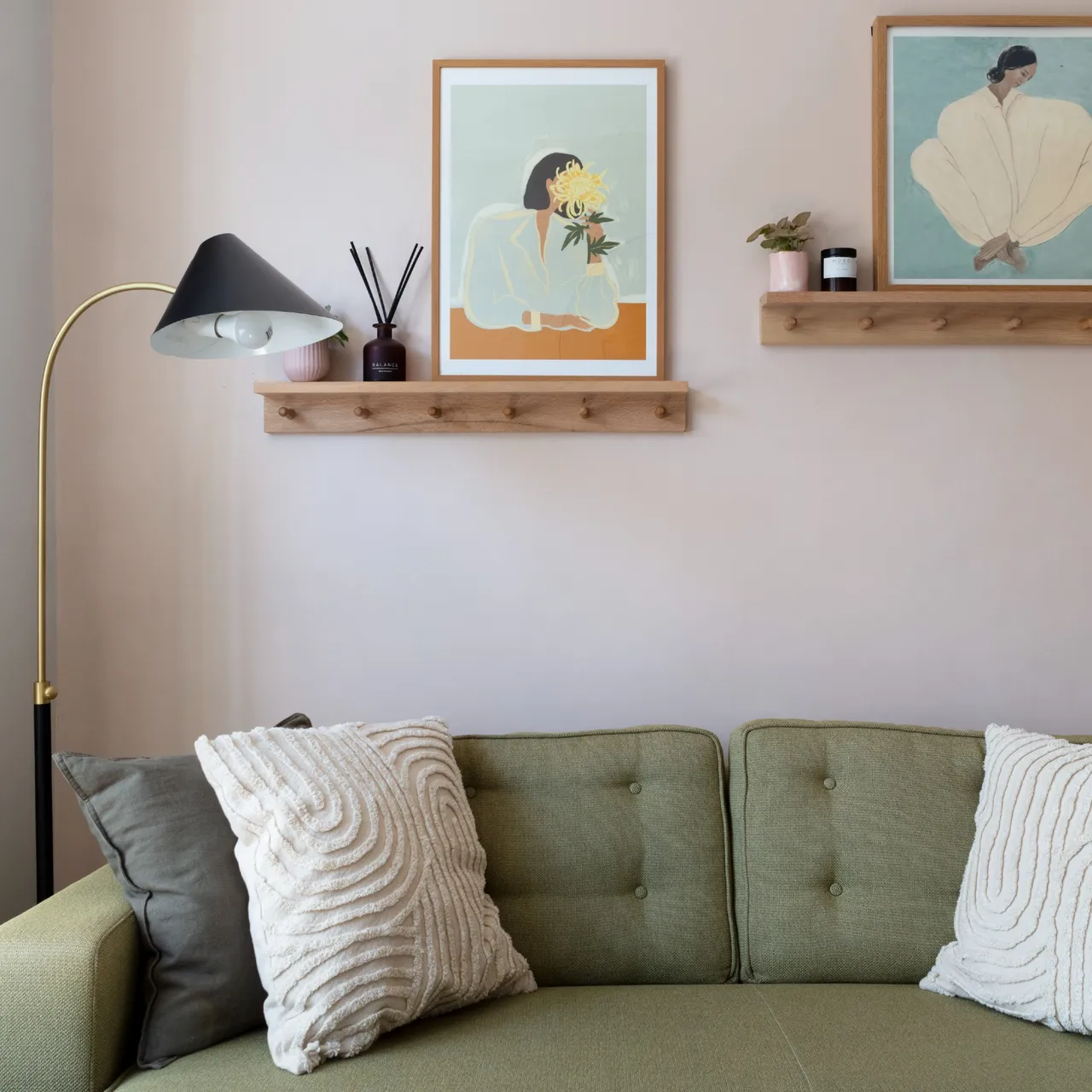 A compact muted green buttoned sofa has pale textured cushions beneath wooden picture shelves