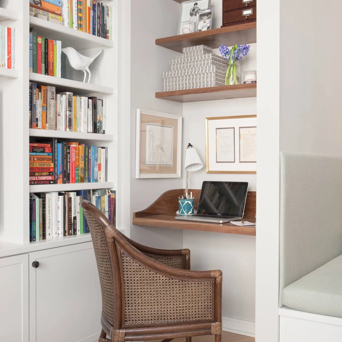 Compact wood desk nook beside white bookshelves with a woven chair, floating shelves and small desk lamp
