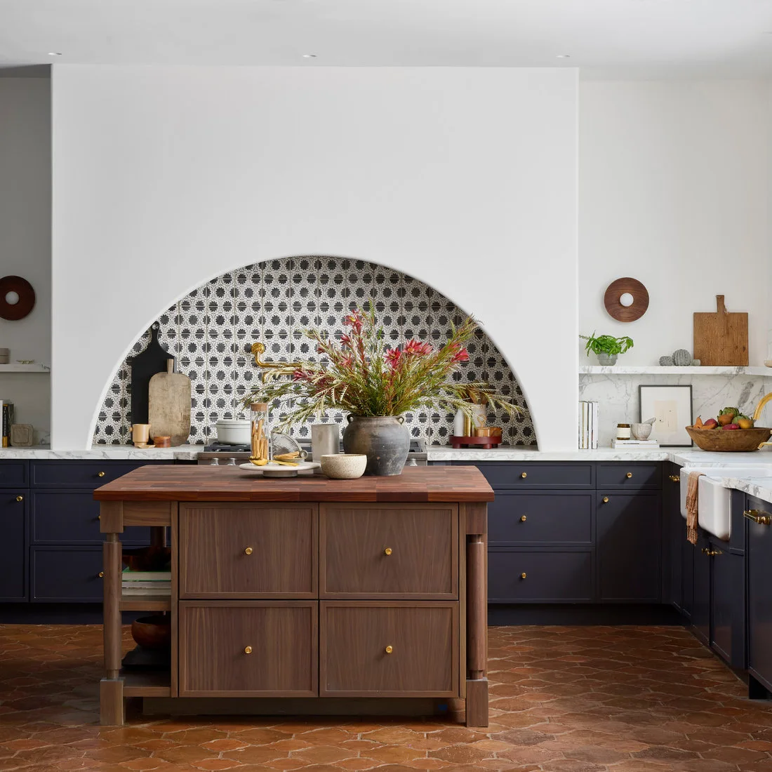 A curved white cooking niche contains patterned tiles above dark cabinets and a timber island on an earthy floor.