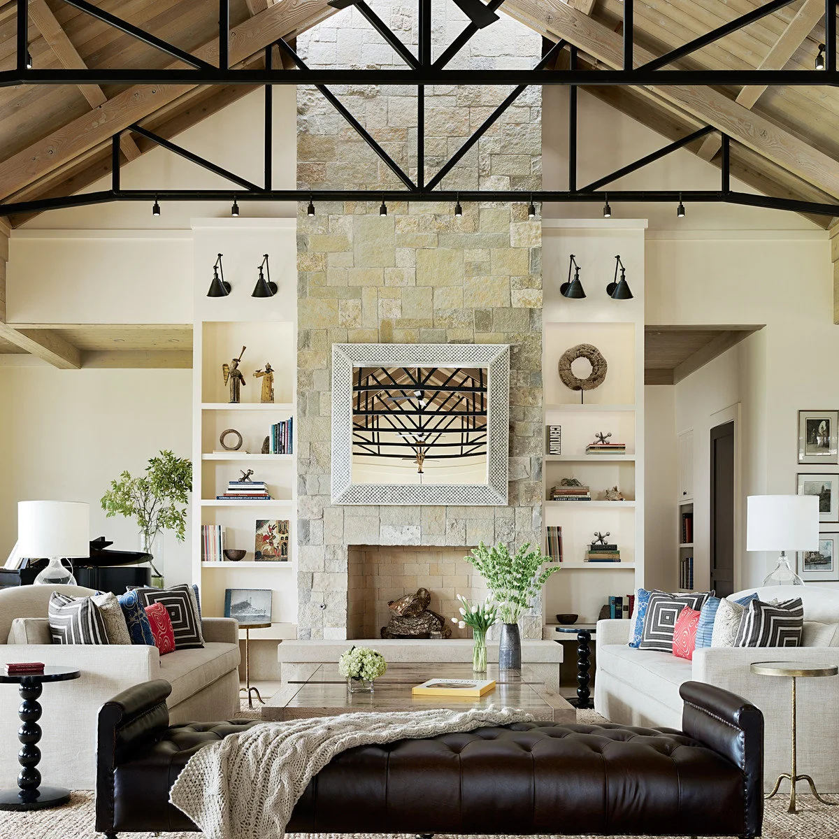 A vaulted living room with black roof trusses, a tall stone fireplace, pale sofas and a dark tufted bench.