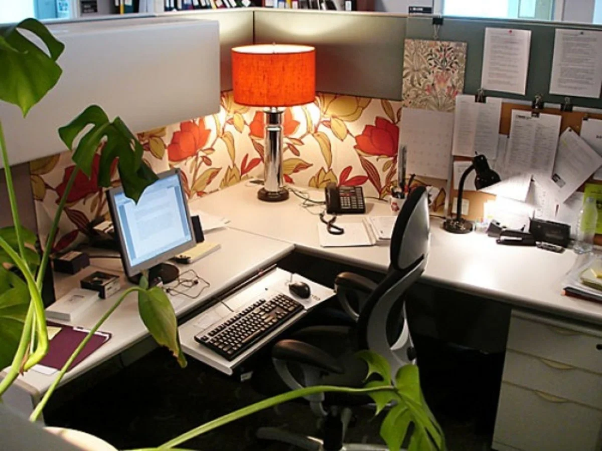Orange floral panel covering behind an L-shaped cubicle desk with an orange-shaded lamp, black task lamp and large green leaves.