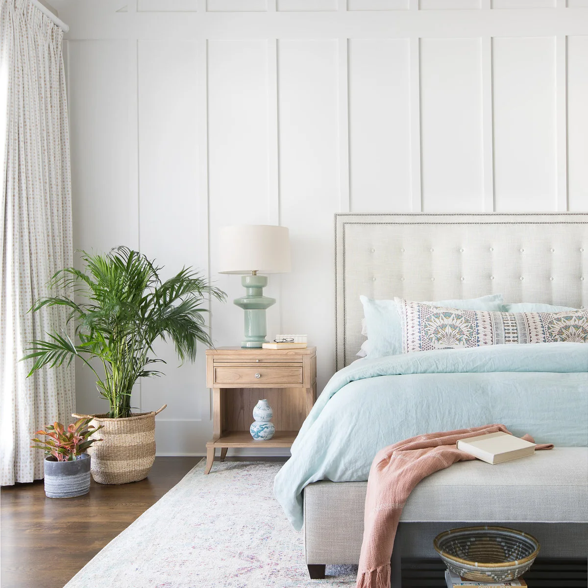 Bedroom with white paneled walls, a cream headboard, pale blue bedding, blush throw and mint bedside lamp