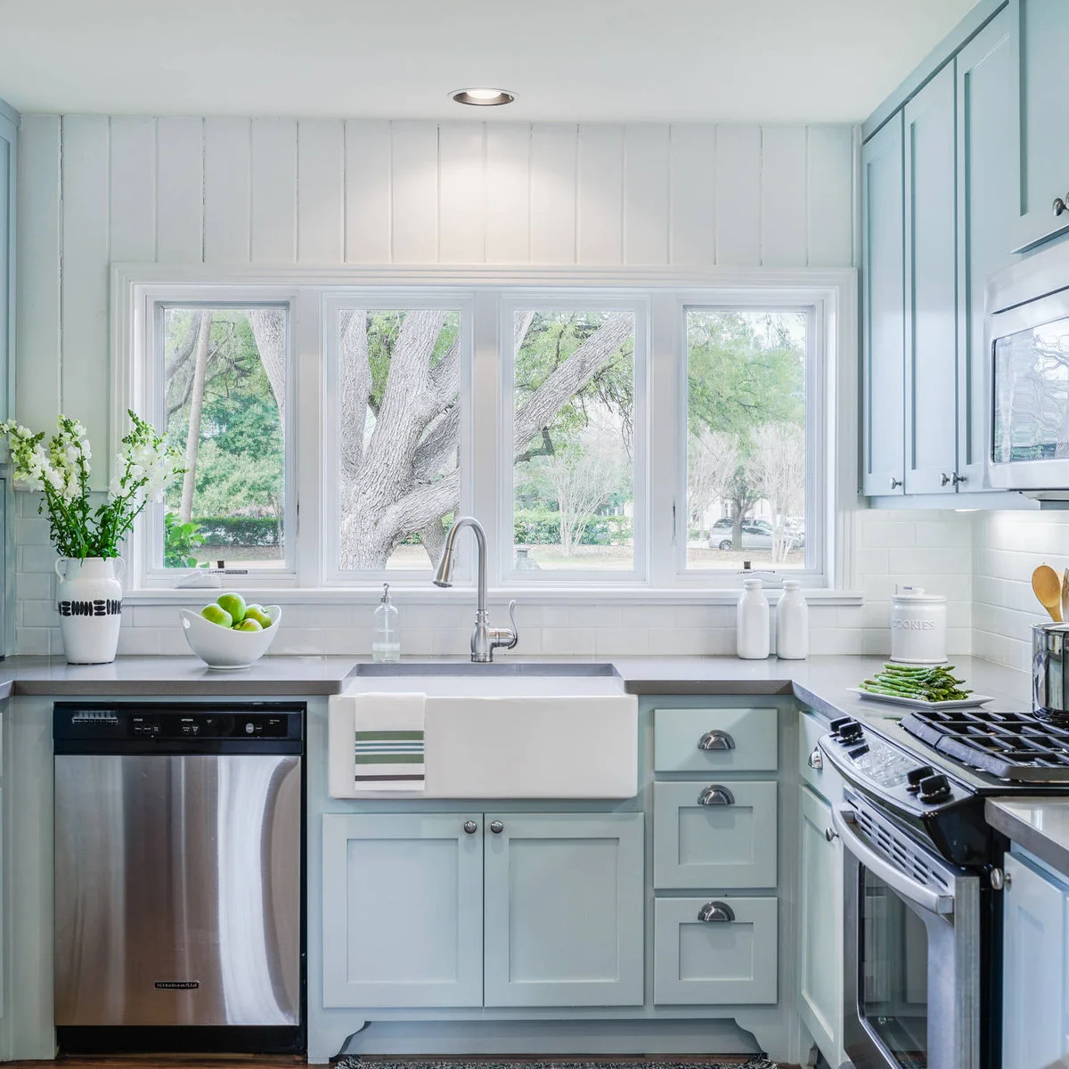 A wide white-trimmed window above a white apron-front sink in a pale blue kitchen.