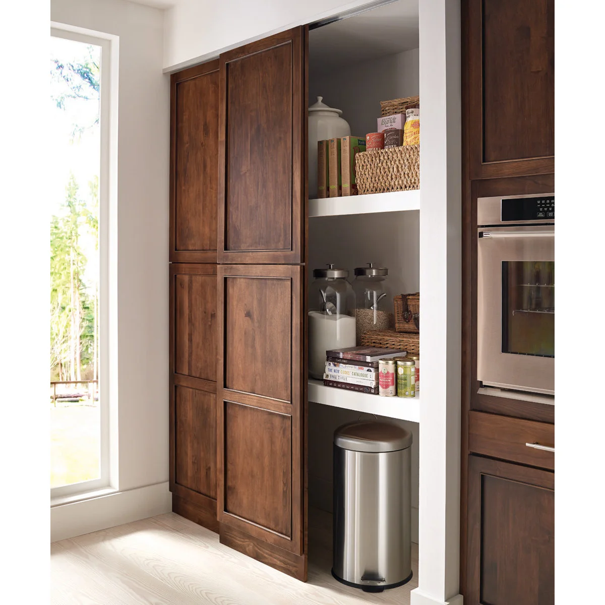 Tall wood panels partly cover white pantry shelves holding jars, baskets, books and a bin beside a built-in oven.