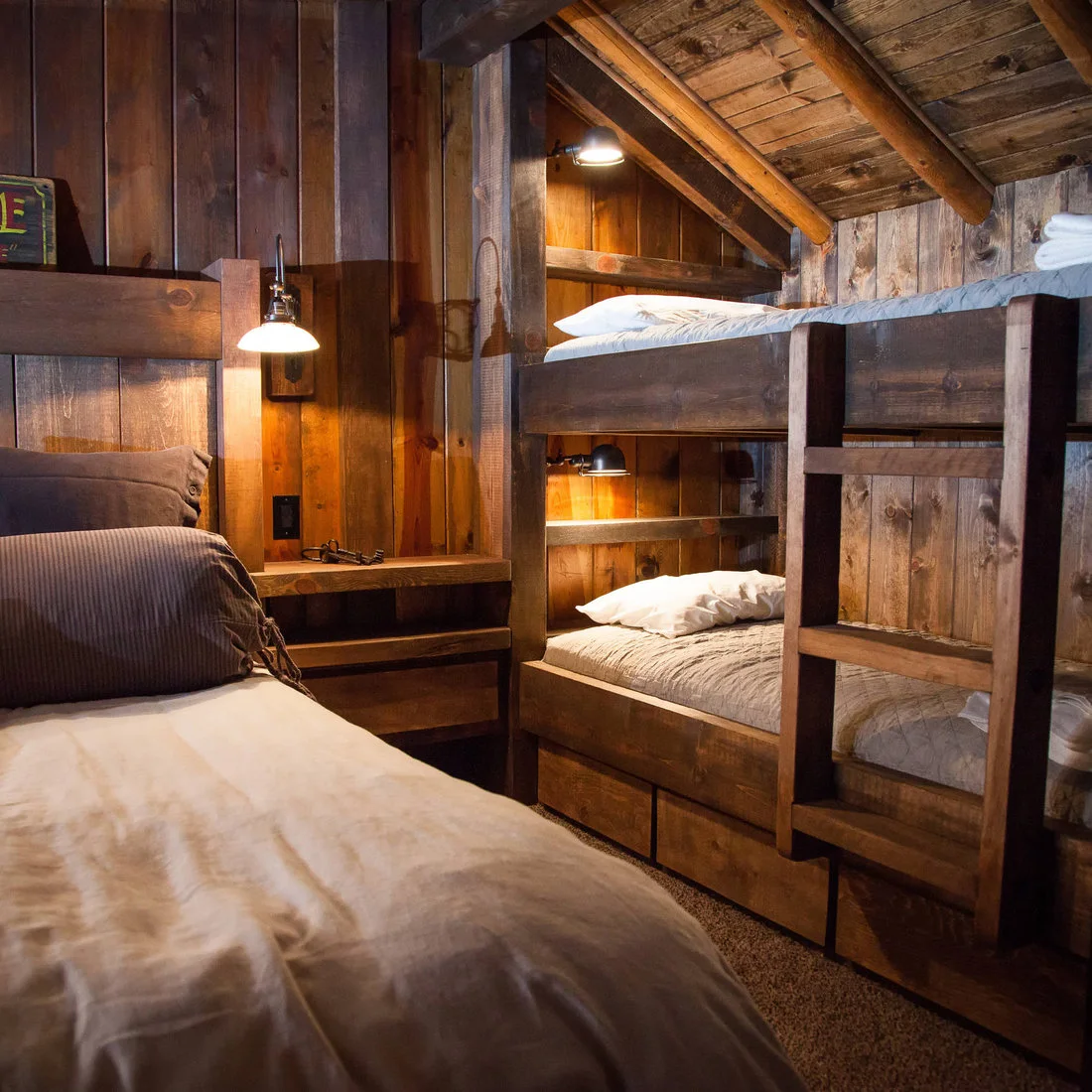 Dark timber beds and built in sleeping bunks with individual wall lamps in a boarded cabin room