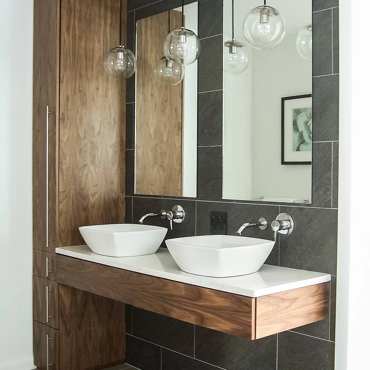 Two white rounded-square vessel sinks sit on a floating wood vanity beside a tall matching cabinet against dark rectangular tiles.