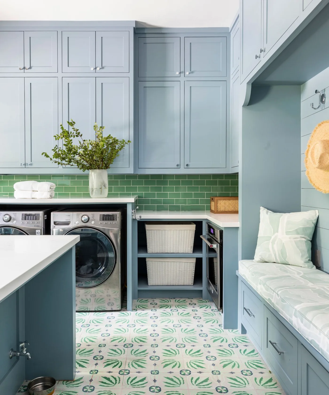 Pale blue cabinets surround laundry appliances, baskets and a cushioned bench beneath hooks, with green wall tiles and a patterned floor.