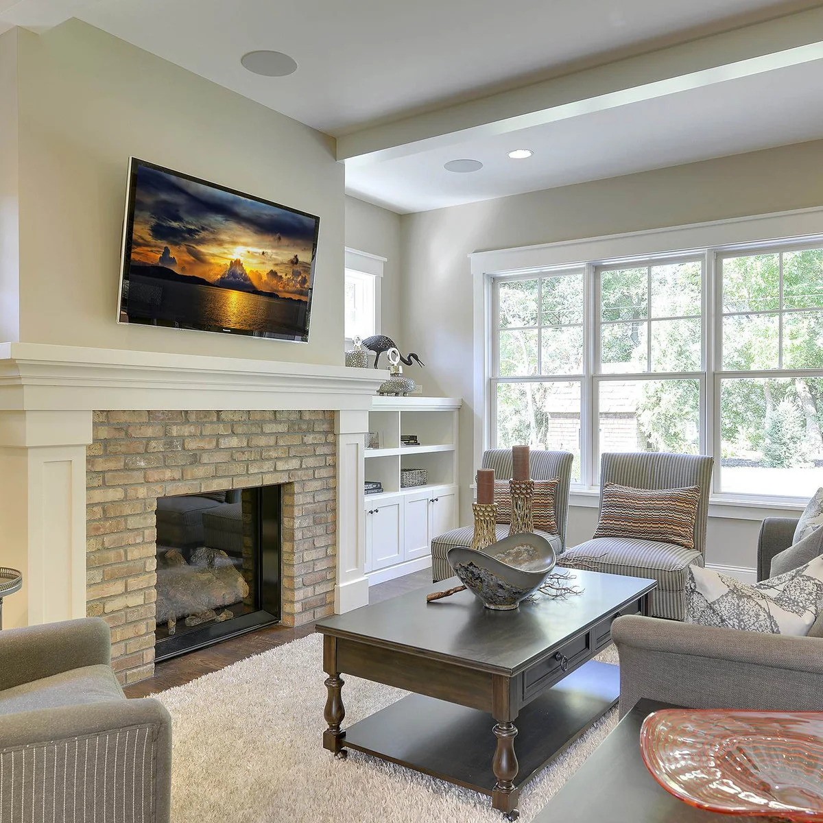 A television above a pale brick fireplace beside windows, with striped chairs, a gray sofa and a dark coffee table.