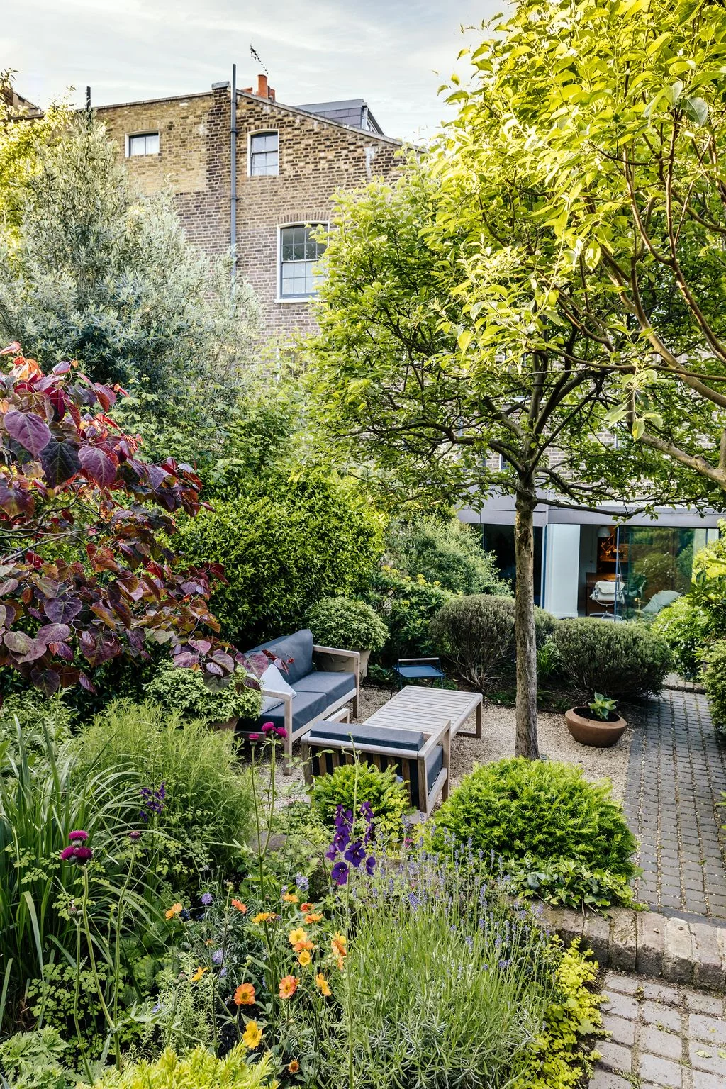 A paved route passes beside a gray outdoor sofa in a leafy compact garden