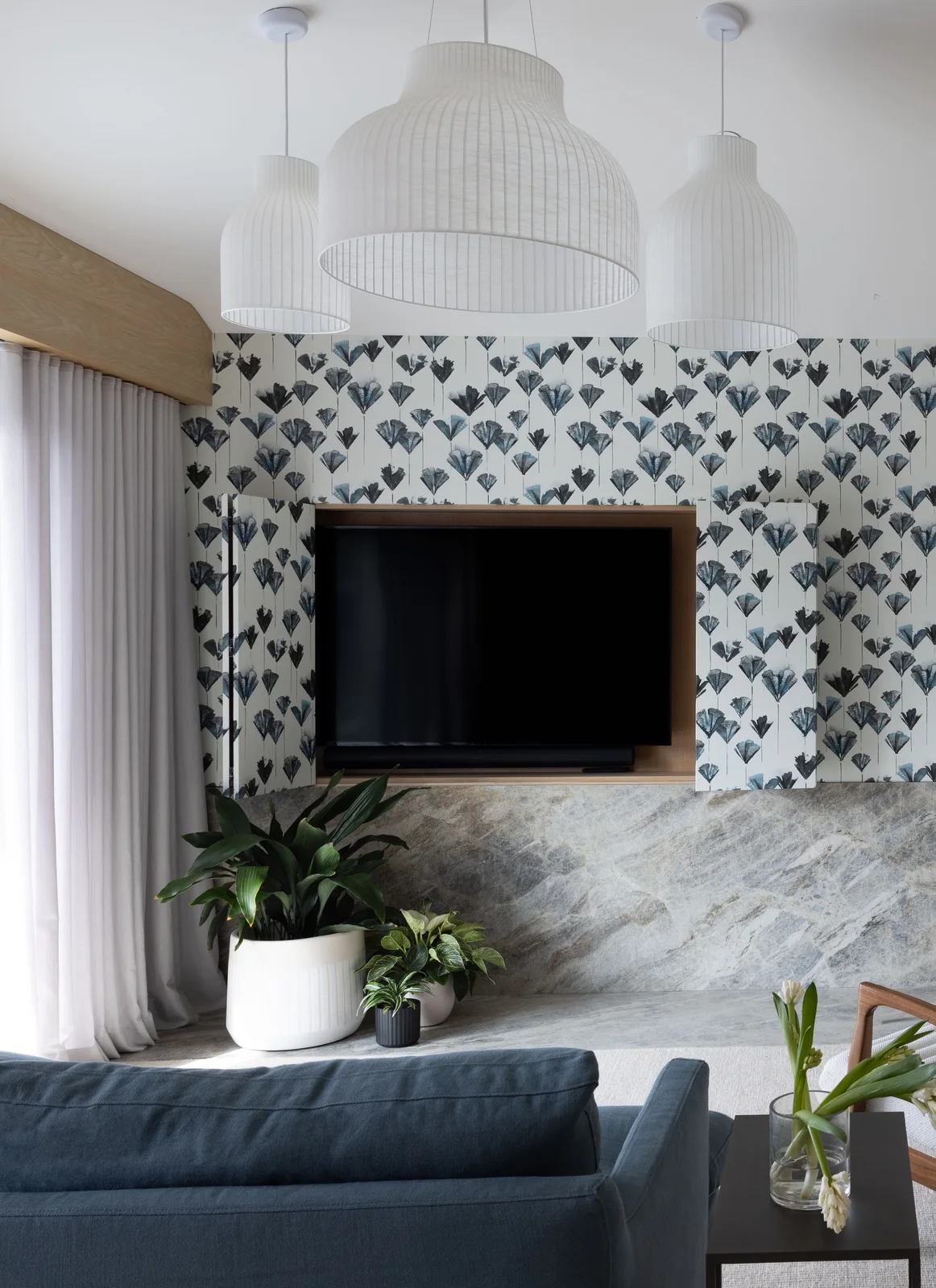 A small blue-and-black leaf wallpaper covers a media wall and open cupboard doors around a television, above a gray veined ledge and blue seating.