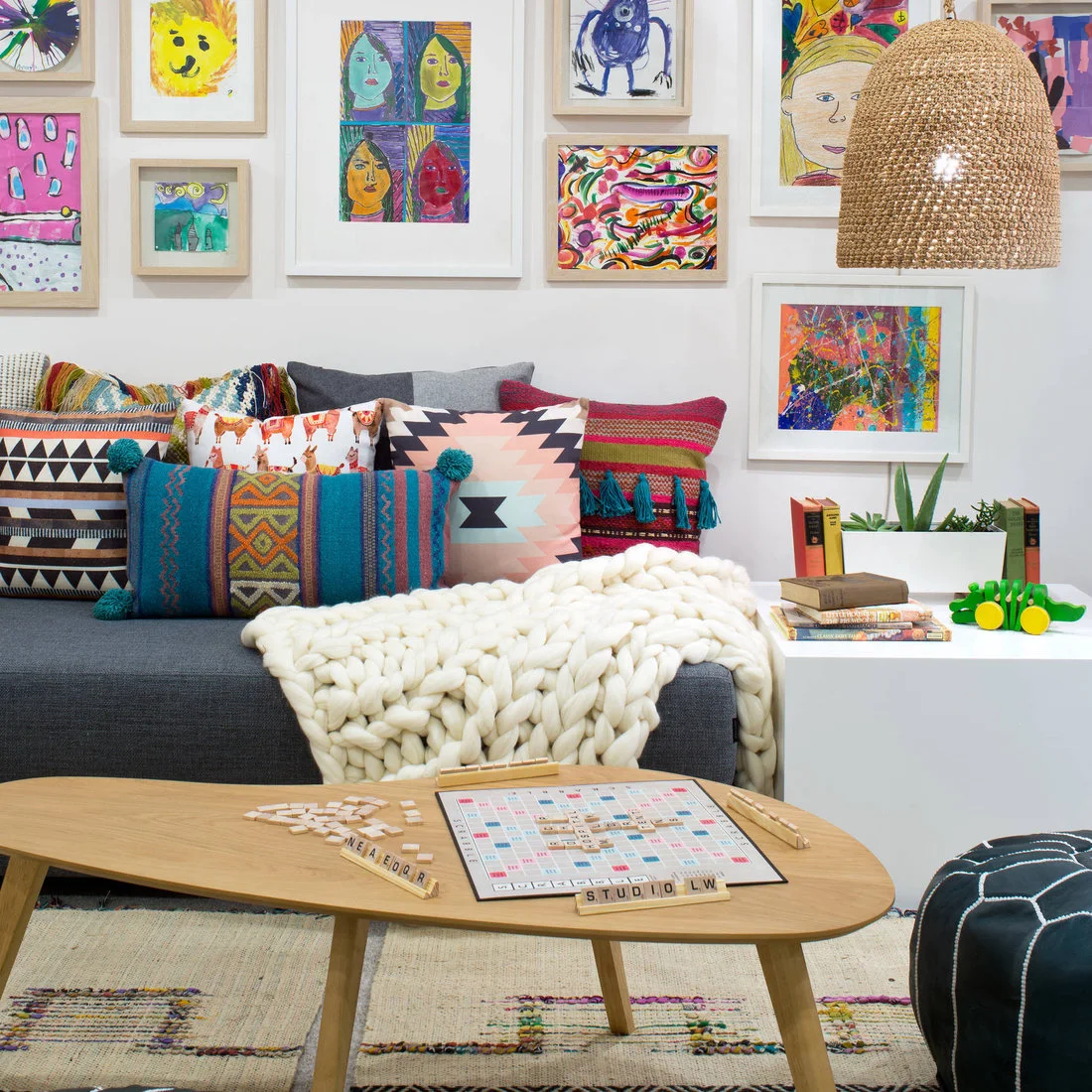 A gray bench with colorful cushions sits beneath framed children’s artwork, beside a low wood table holding a board game and a woven pendant shade.