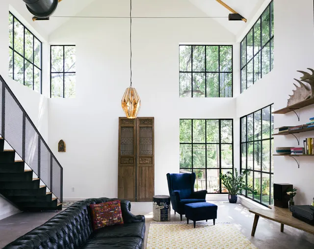 A black tufted sofa sits near broad glazing and pale walls in a tall room.
