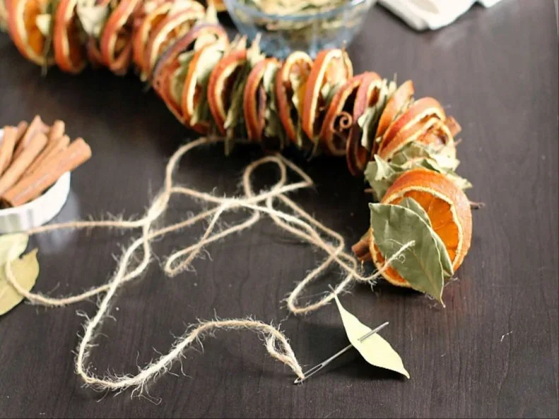 Dried orange slices and pale leaf shapes are threaded along cord on a dark work surface beside cinnamon-like sticks