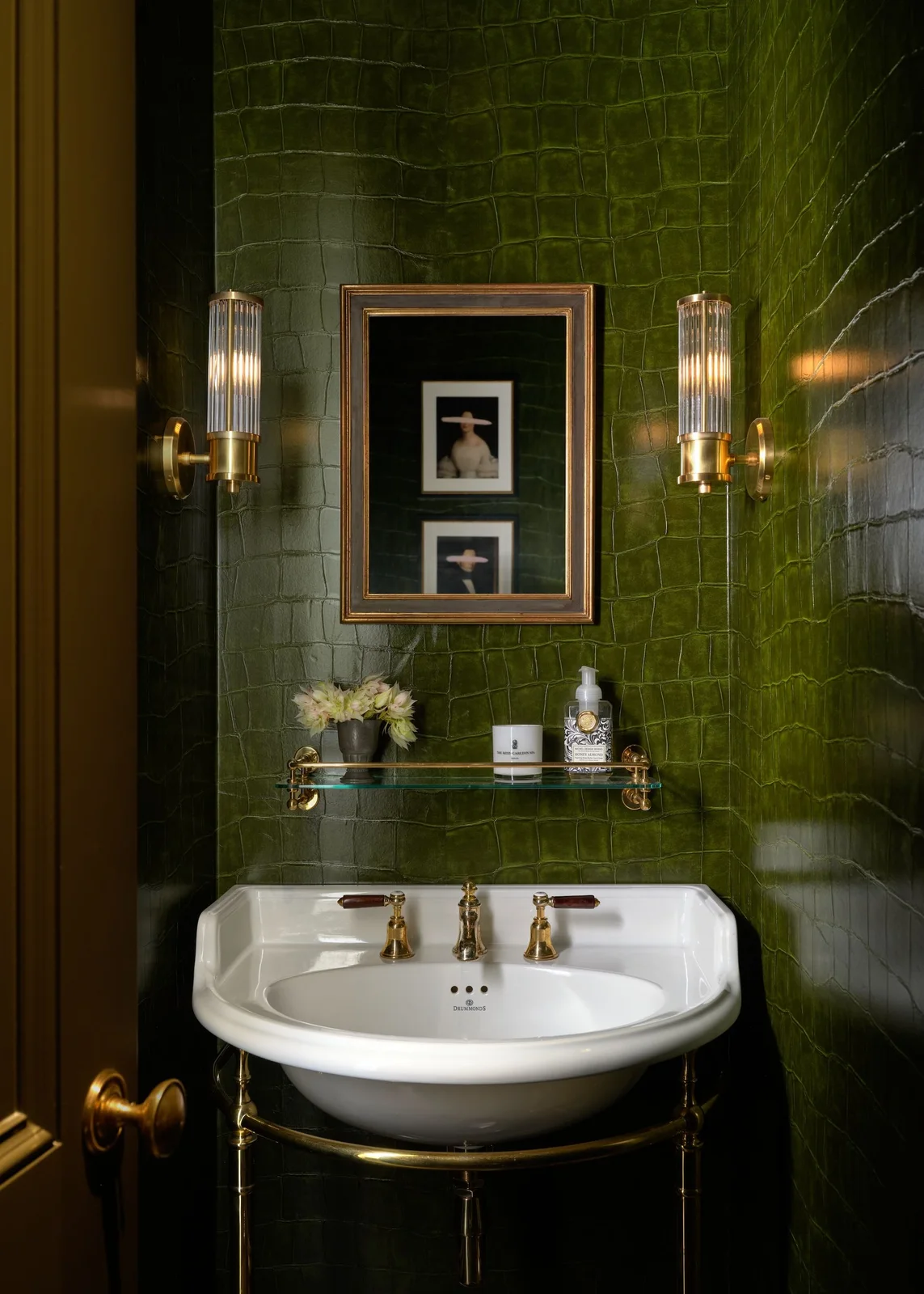 Dark crocodile-effect olive wallpaper surrounds a white console basin with brass fittings, a framed mirror and two ribbed wall lights.