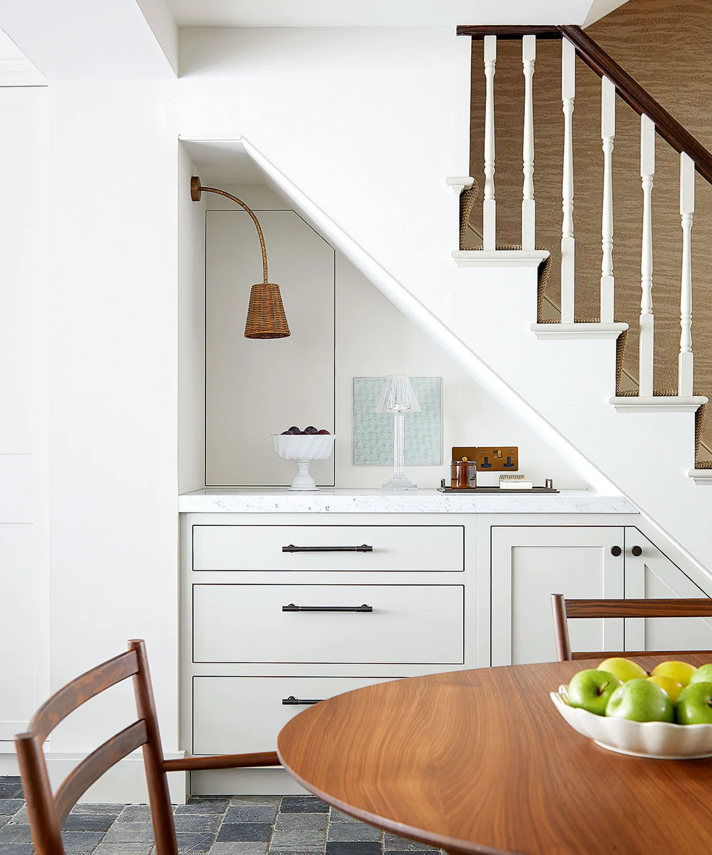 White drawers and cupboards beneath stairs beside a wood dining table, mirror and two lamps