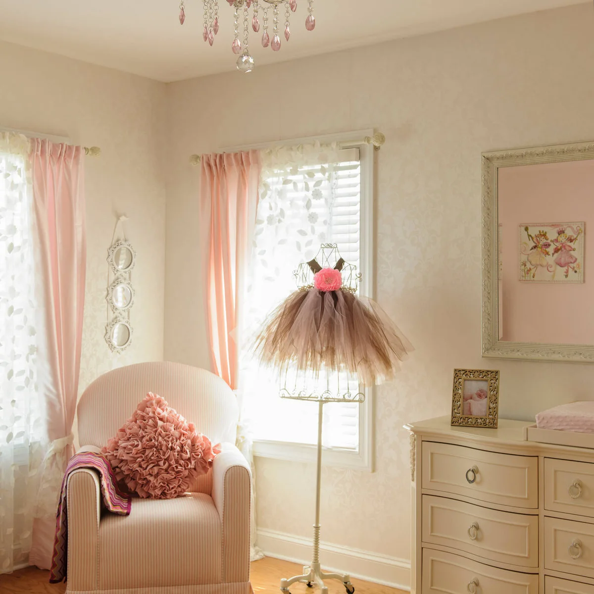 Nursery corner with a pale striped chair, pink curtains, cream dresser and framed pink artwork