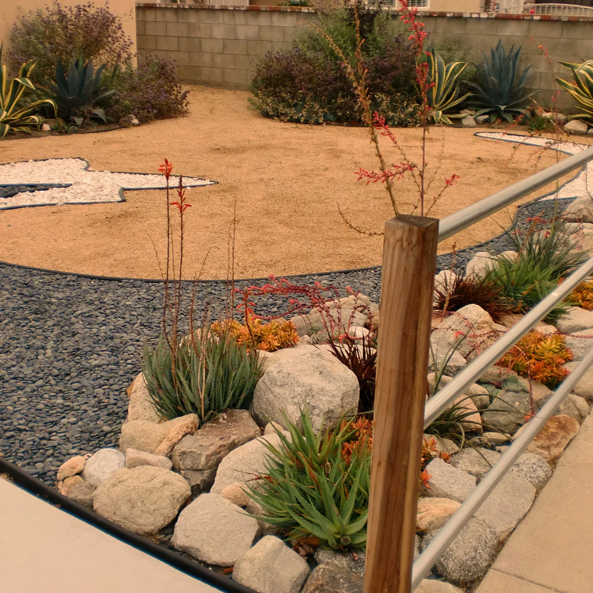 Slim horizontal metal rails between wood posts beside a gravel garden with boulders and flowering succulents.