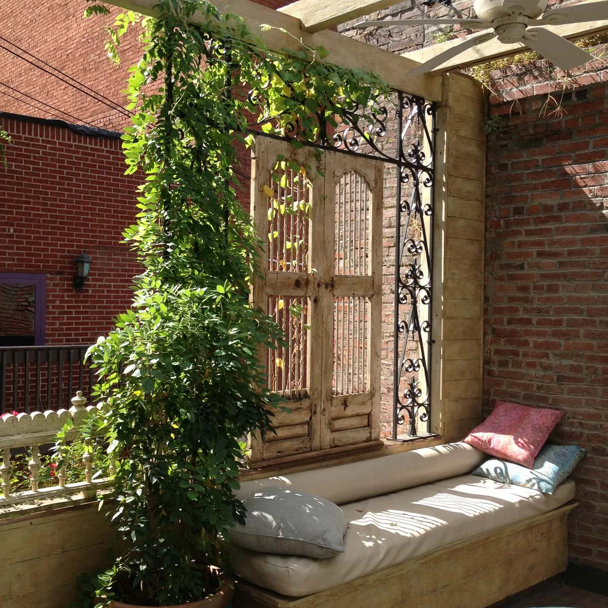 A pale cushioned wood daybed sits against brick beneath a pergola, beside climbing greenery and decorative wood doors.
