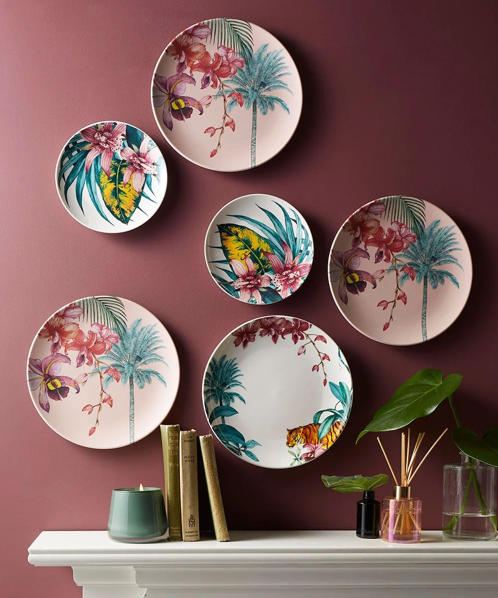 Six decorative floral plates in a loose grouping on a pink-plum wall above a pale ledge holding cups, books, a plant, and a diffuser.