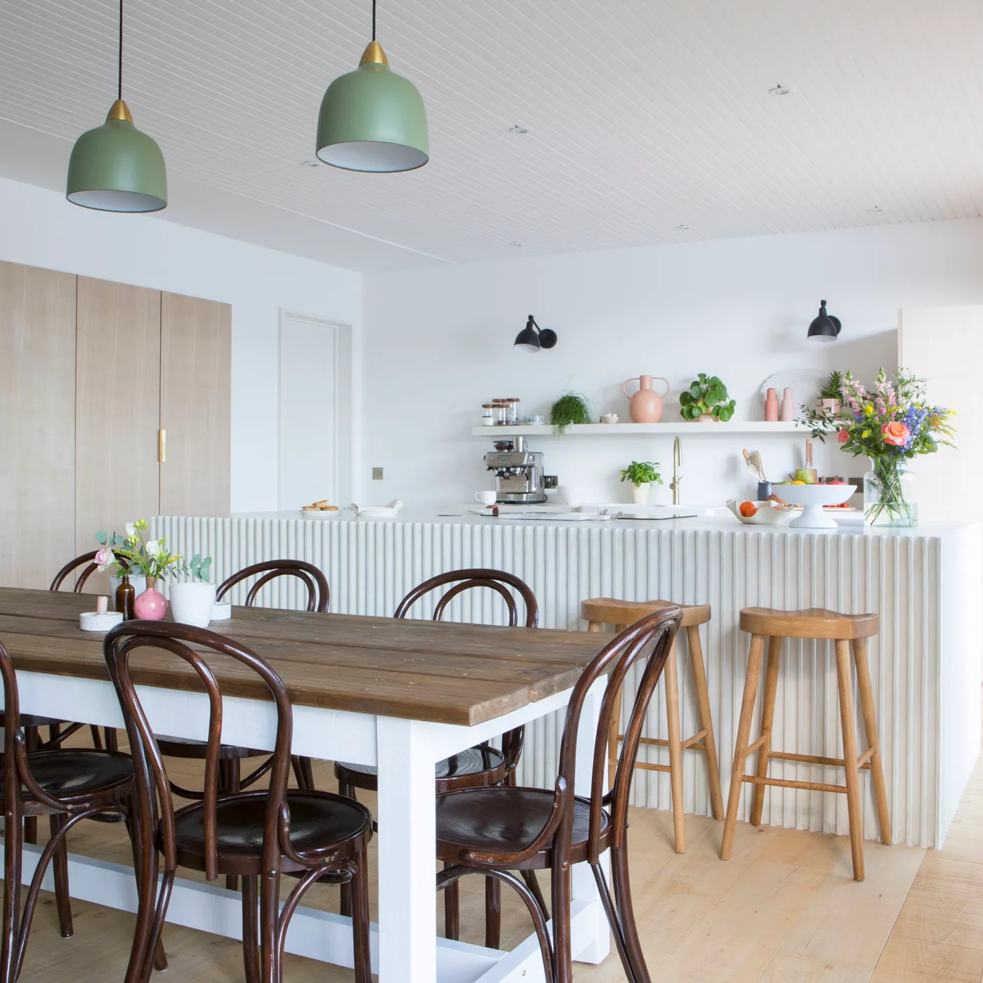 Open kitchen and dining space with a wooden table, dark curved chairs, pale island, green pendant shades and warm-toned flooring.