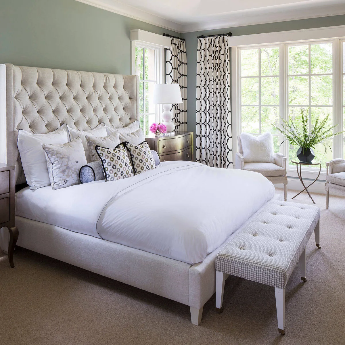 A pale upholstered bed with a bench at its foot, plus two small chairs and a round table beside a large window.