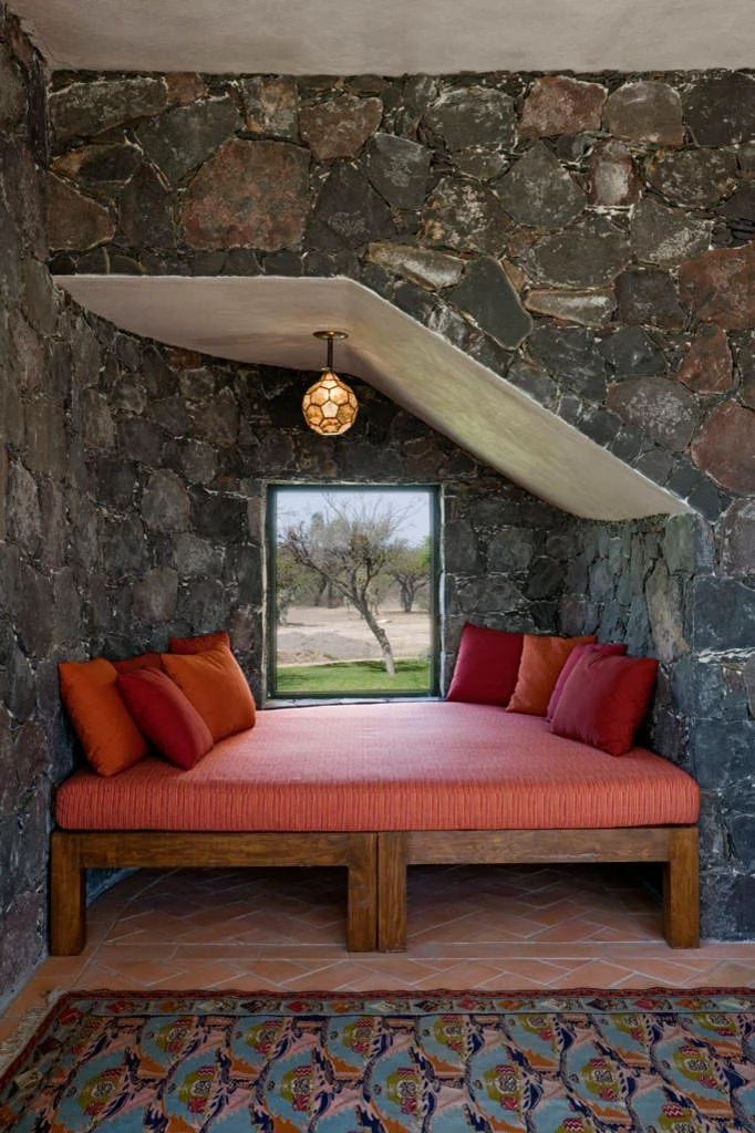 Orange cushioned wood daybed with red and orange pillows in a stone-walled recess beneath a sloping stair underside.
