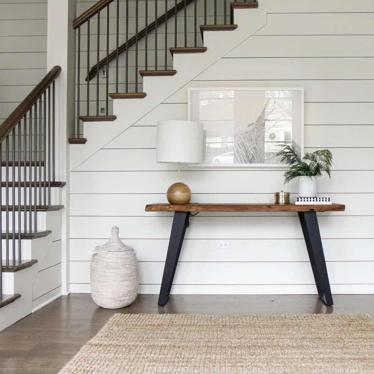 A wood-topped console beneath staircase paneling, with a rounded lamp on one side and books and greenery on the other.