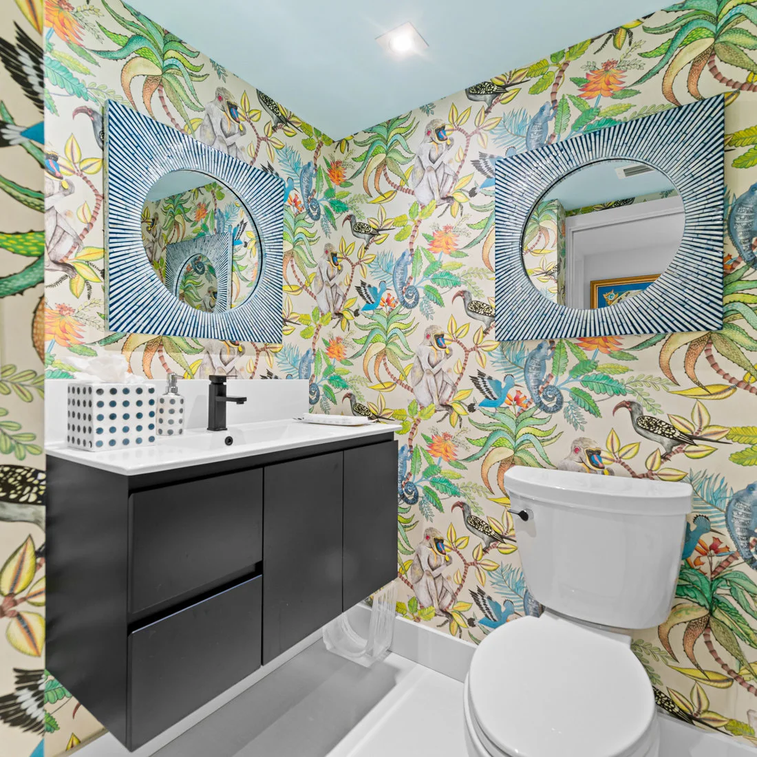 Colorful botanical and animal wallpaper surrounds two blue radial mirror frames above a dark floating powder-room vanity and white toilet