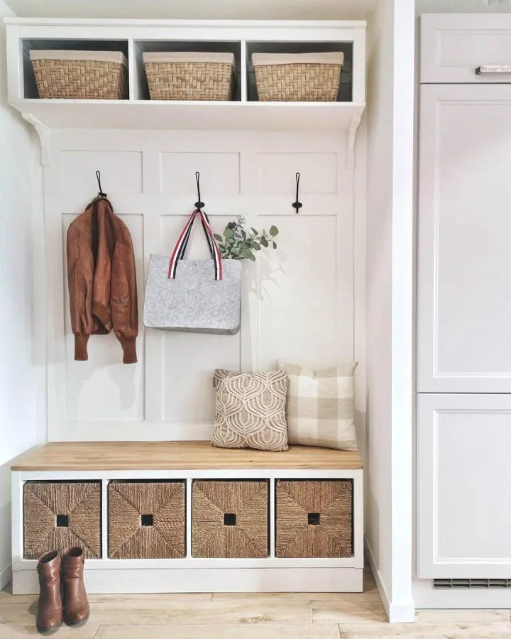 White entryway unit with wood-topped lower cubbies, four woven baskets, black hooks and three upper baskets beside a tall cabinet.