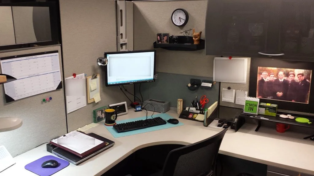 Gray cubicle with an elevated monitor, pale blue keyboard mat, wall calendar, small shelf and a separate screen on a raised side stand.
