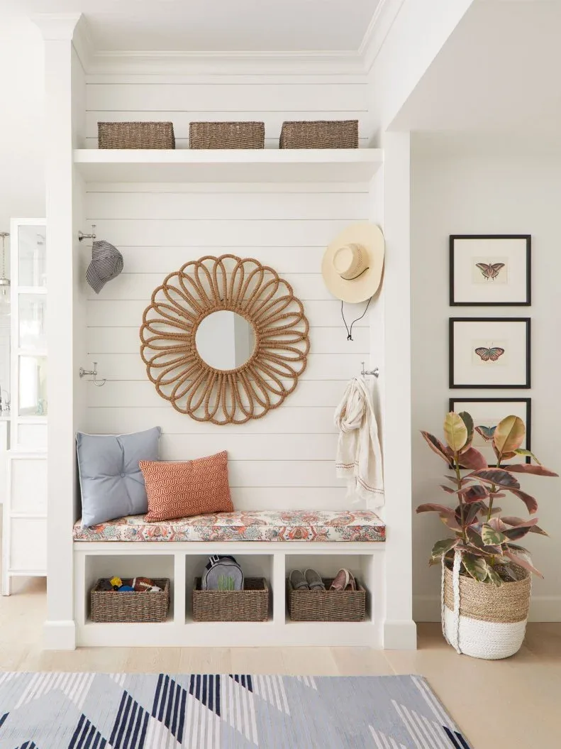 White paneled mudroom bench with woven baskets, looped round mirror, patterned cushion, small pictures and potted plant.