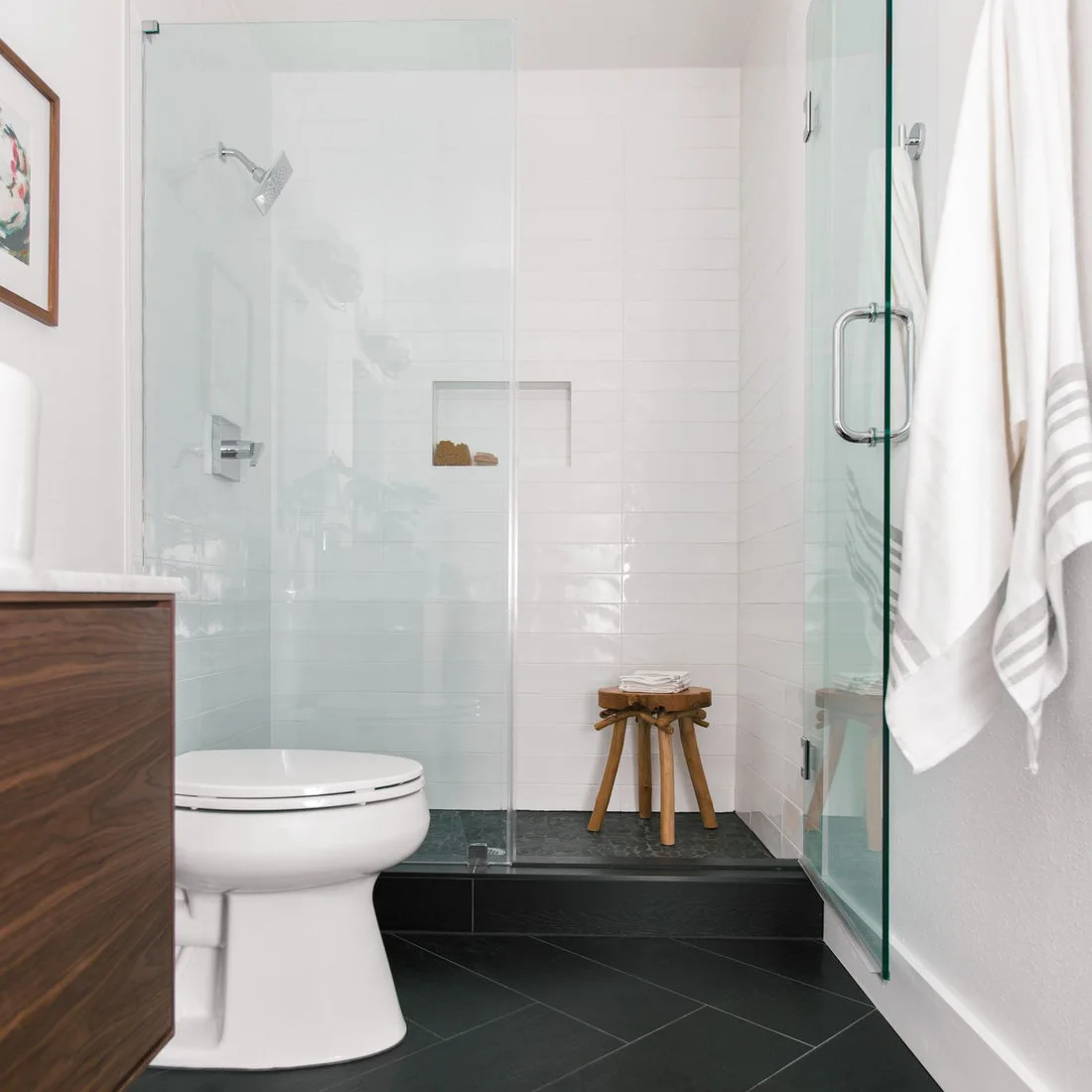 A narrow bathroom has a wood vanity edge, white toilet and clear shower enclosure above a dark tiled floor.