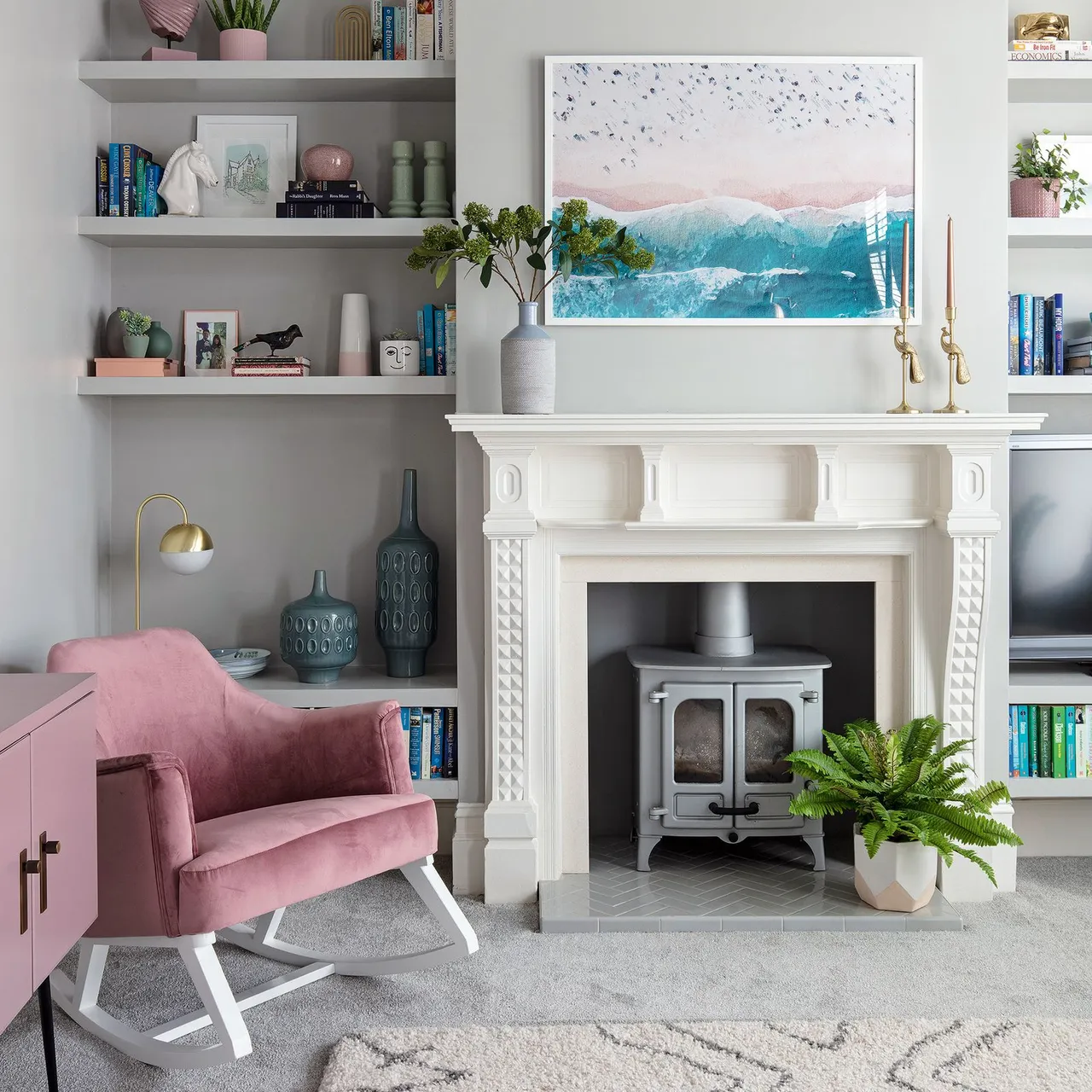 A pale carved fireplace surround frames a gray stove beside a pink rocking chair and fitted shelves.