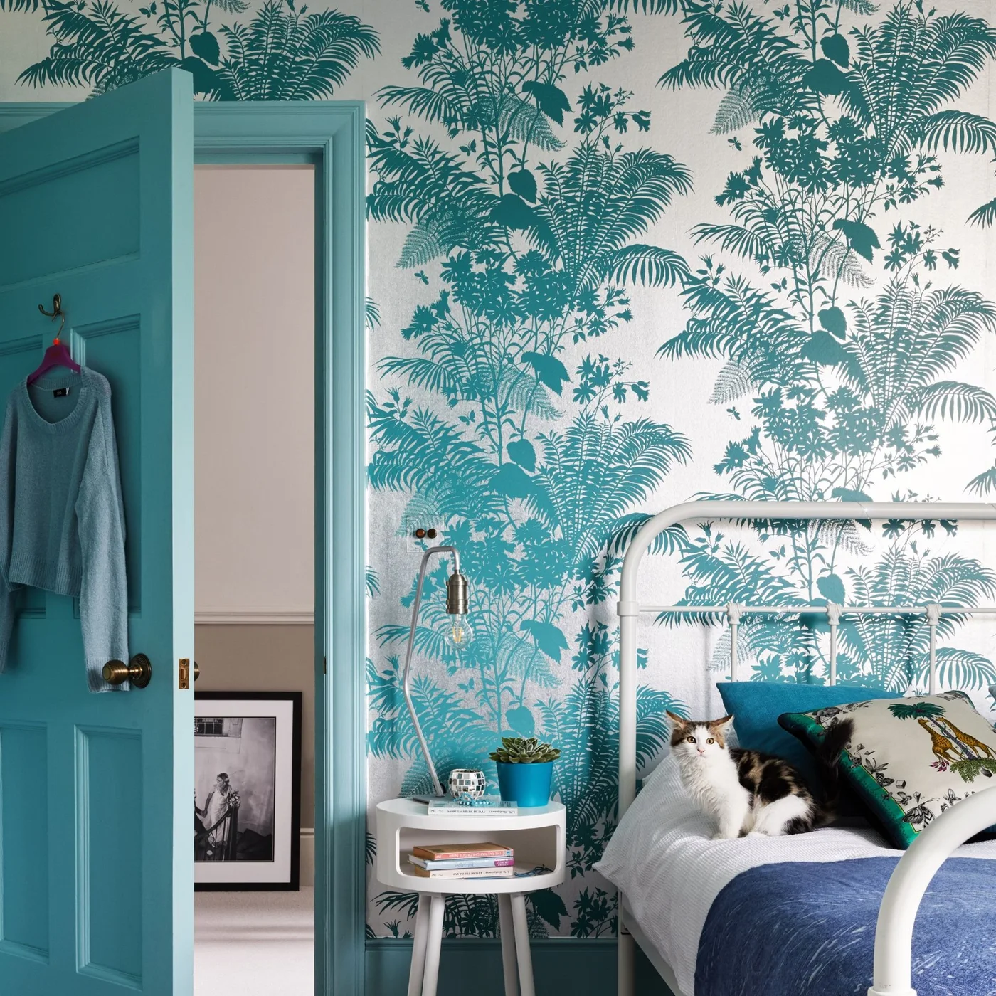 Teal tree and palm silhouettes on pale bedroom wallpaper connect with a teal door beside a pale metal headboard and blue bedding