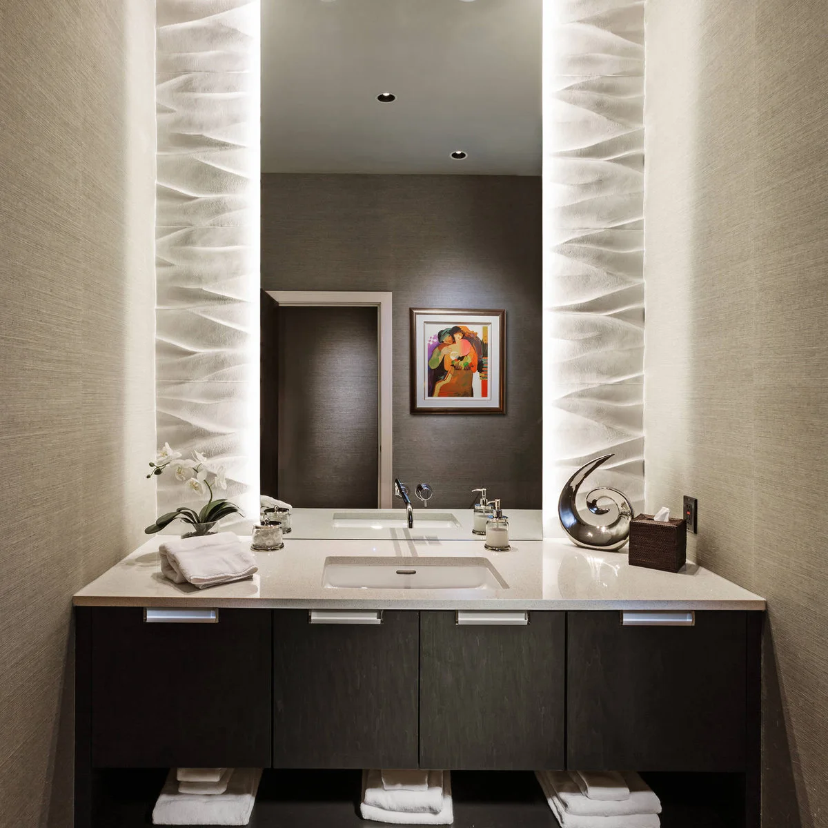 A broad dark vanity beneath a tall mirror flanked by illuminated sculptural pale wall panels.