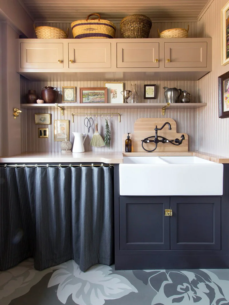 Four differently shaped woven baskets above a muted kitchen wall cabinet with a decorated shelf and sink below.