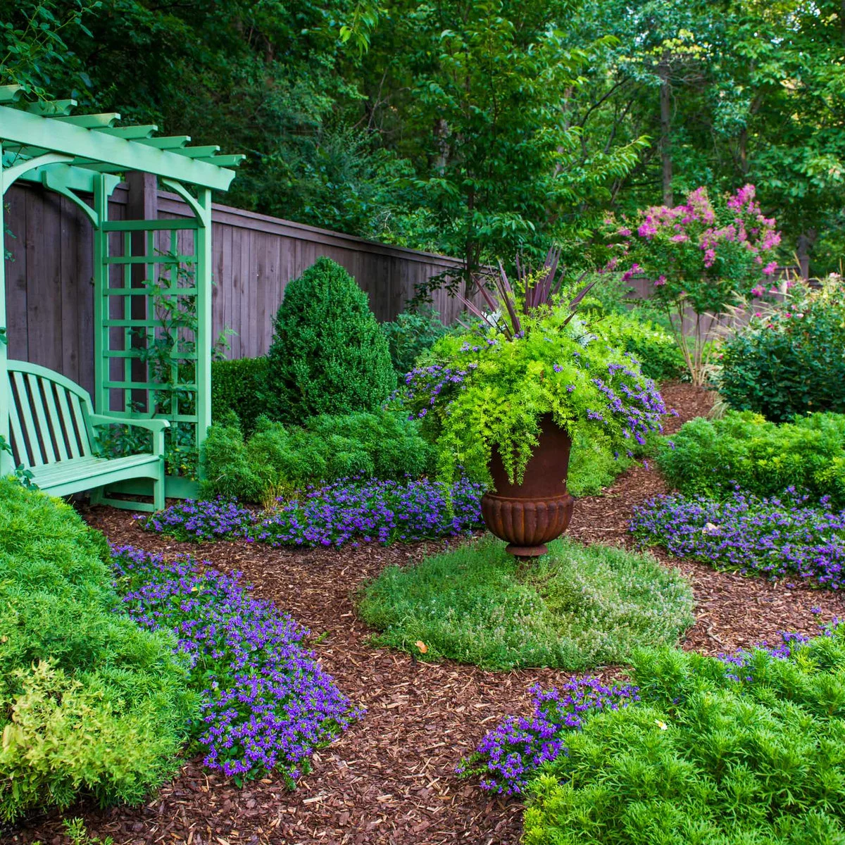 A pale green arbor shelters a bench beside a dark fence, with purple flowers and a tall planted urn nearby.