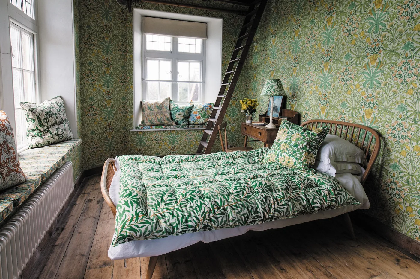 Green and yellow floral wallpaper around a wooden bed and window seat with green botanical bedding.