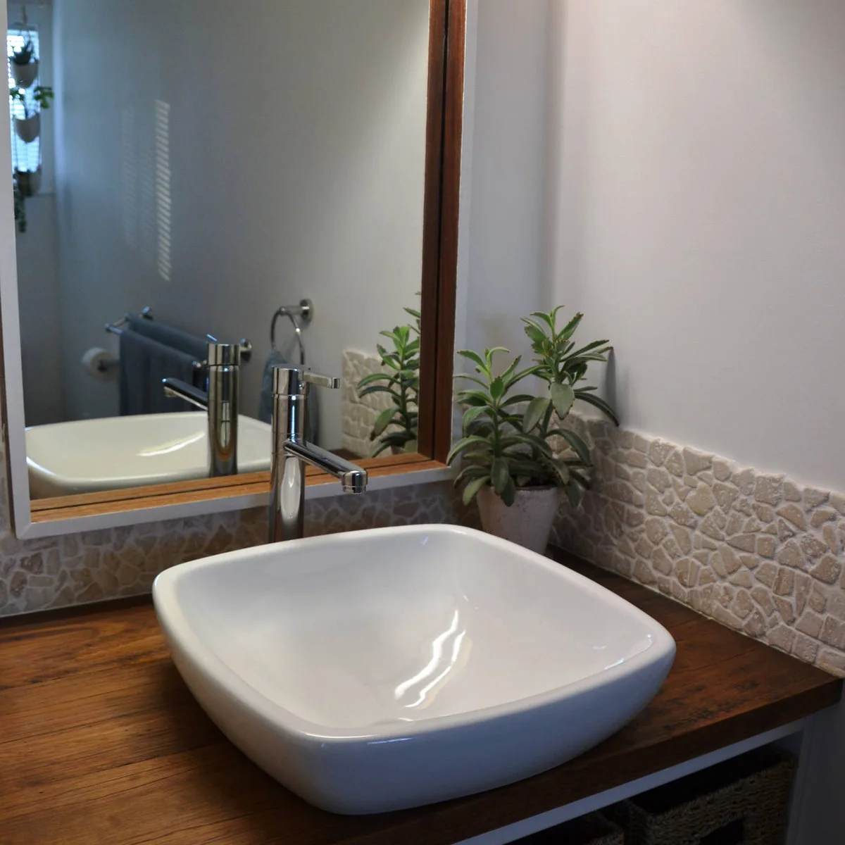 White vessel basin and tall chrome faucet on a wood counter with a low pebble-pattern backsplash and framed mirror.