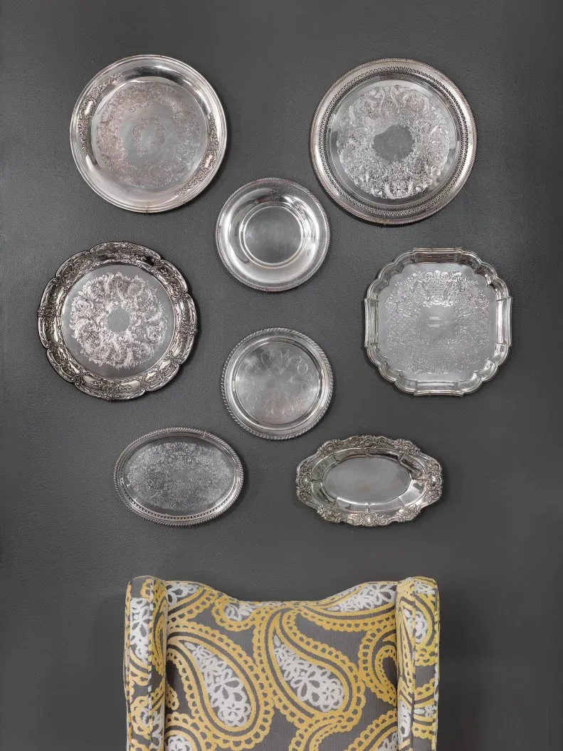 Cluster of silver-toned round, oval, and rectangular decorative trays on a charcoal wall above a partially visible yellow-and-gray patterned chair.