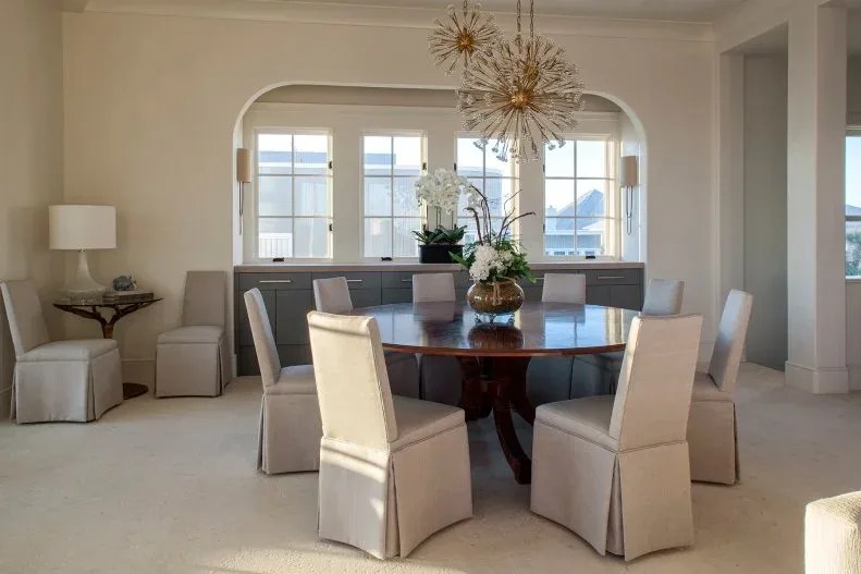 Pale covered chairs around a round wooden table beneath two gold-toned starburst lights beside an arched opening and windows.