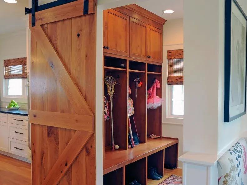 A wood bench with open shoe cubbies sits below tall coat bays and closed upper cupboards beside a sliding wood door.