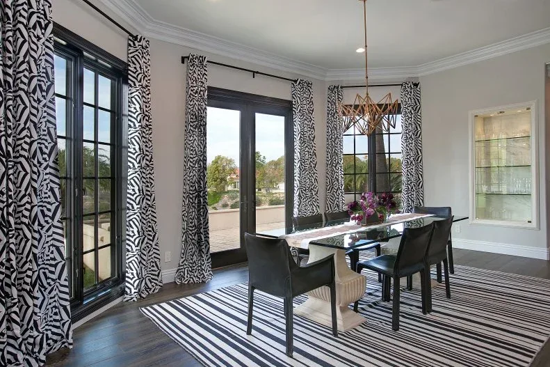 Dining room with black glazed doors, patterned black-and-white curtains, striped rug, pale-topped table and dark molded chairs