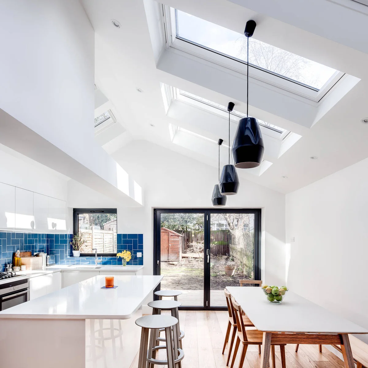Terraced-house kitchen with a white breakfast island, dining table and blue tiled cooking wall beneath rooflights